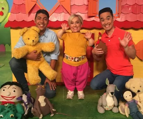 Two men and a woman with short hair wave cheerfully in a colorful setting with plush toys.