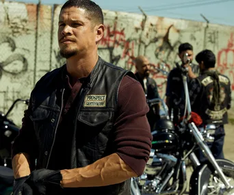 "Man in leather vest labeled 'Prospect' stands by motorcycles with fellow bikers in the background, graffiti-covered wall."
