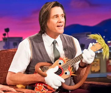 Man sitting on a talk show set, holding a colorful puppet-like ukulele, with a vibrant background.