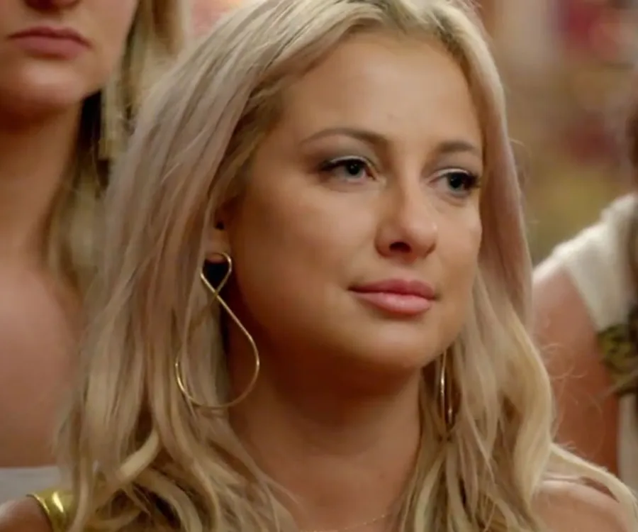 A woman with long blonde hair and large hoop earrings looks pensive while surrounded by others in a room.