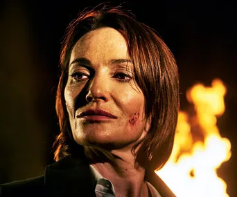 Actor with a cut on cheek in front of fire, intense expression, wearing dark clothing, from "Bancroft" series.