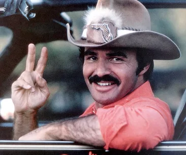 Man in cowboy hat smiling and giving a peace sign from a car window.