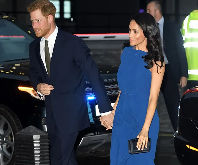 Couple holding hands, the woman in a blue dress and the man in a dark suit, walking near parked cars.