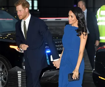 Couple holding hands, the woman in a blue dress and the man in a dark suit, walking near parked cars.