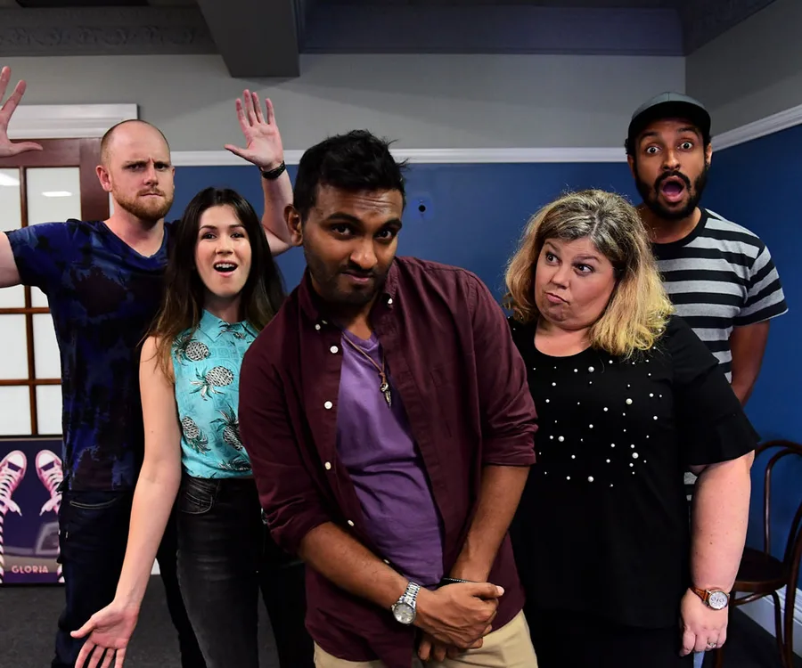 Comedy show cast posing playfully with various expressions against a blue wall.