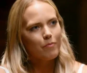A woman with long blonde hair appears thoughtful, wearing a light-colored top, against a blurred dark background.