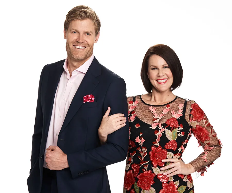 Man in a dark suit with a pink shirt and pocket square, standing beside a woman in a floral dress, both smiling.