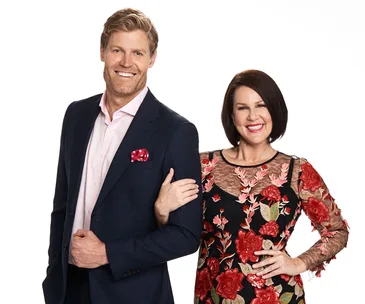 Man in a dark suit with a pink shirt and pocket square, standing beside a woman in a floral dress, both smiling.