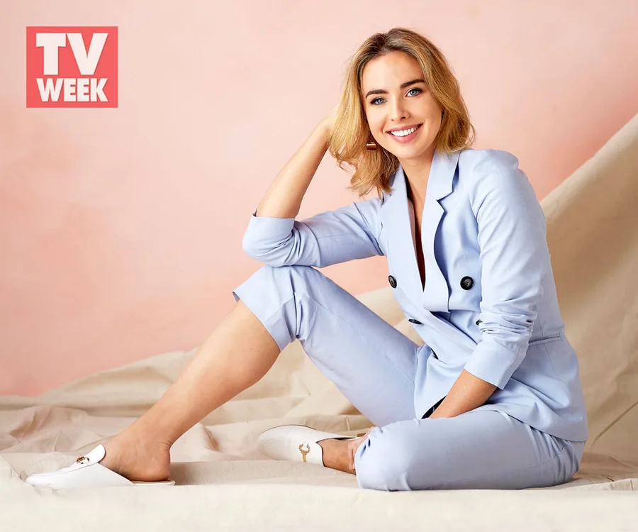 Brightly dressed woman in a light blue suit sitting on the floor, smiling, with a backdrop featuring a TV Week logo.