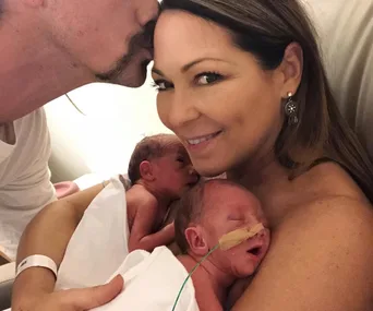 Family with newborn twins; woman holding babies, partner kisses her forehead.