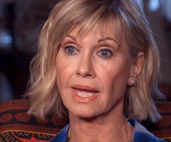 A woman with short blonde hair speaks during an interview, with a patterned background.