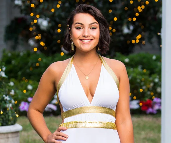 Woman in a white and gold dress poses outdoors with sparkling lights and greenery in the background.