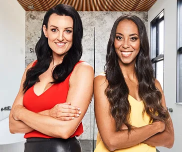 Two women smiling with arms crossed, one in a red top and the other in yellow, standing in a modern room with marble walls.