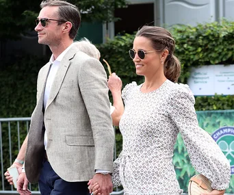 Couple walking outside at an event, holding hands, both wearing sunglasses; woman in a patterned dress, man in a grey suit.