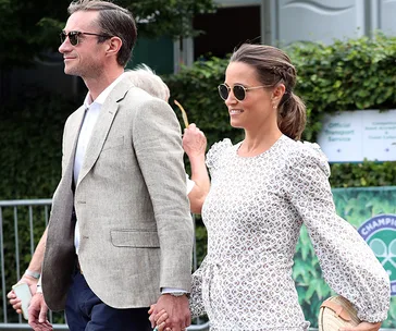 Couple walking outside at an event, holding hands, both wearing sunglasses; woman in a patterned dress, man in a grey suit.