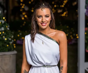 Jamie-Lee in a white one-shoulder dress with gold accents, adorned with a headpiece, smiling at a night event.