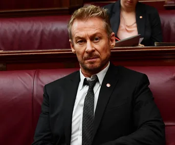 Chefs Cleaver, a character from the series "Rake," sitting in an official setting, wearing a suit and tie.