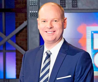 A smiling man in a blue suit and striped tie stands in a brightly lit studio setting.