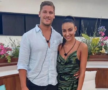 A smiling couple standing close together outdoors, surrounded by plants and flowers.