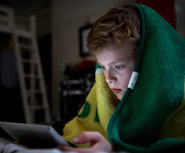 Boy under blanket playing on a tablet in a dimly lit room.