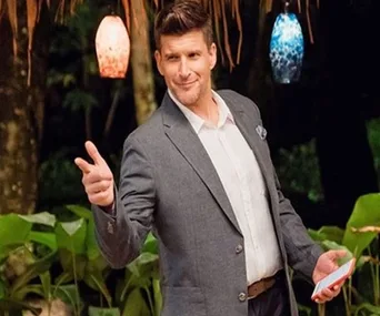 Host in a suit gestures with a playful smile on "Bachelor in Paradise Australia," holding a card, with colorful lamps above.