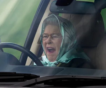 An elderly woman wearing glasses and a headscarf yawns while sitting in a car.