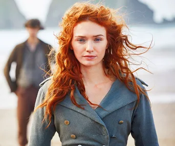 Red-haired woman in blue coat stands on a beach with a blurred figure in the background from a scene in "Poldark."