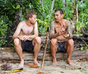 Two shirtless men sitting on a log in a jungle setting, engaged in conversation.
