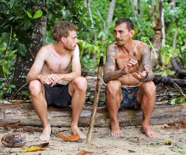 Two shirtless men sitting on a log in a jungle setting, engaged in conversation.