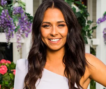 A woman with long brown hair smiles brightly, wearing a white top, standing in front of a floral backdrop.