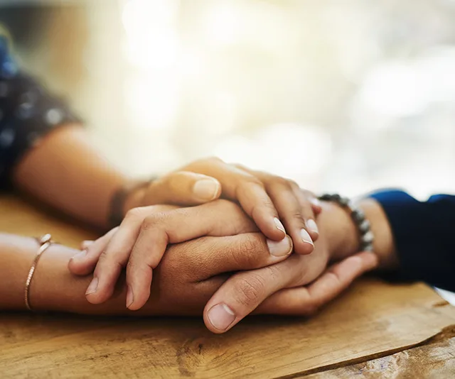 Two people holding hands on a wooden table, symbolizing support and comfort.