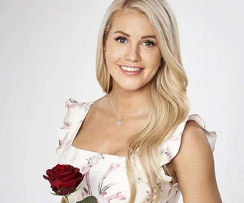 Woman smiling and holding a red rose, wearing a floral dress with long blonde hair.