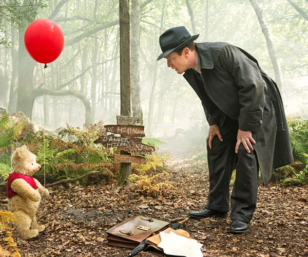 Christopher Robin bends down to speak with Winnie the Pooh holding a red balloon in a forest setting.