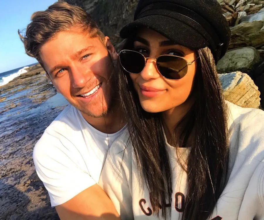 A couple smiles on a rocky beach, the man in a white shirt and the woman in sunglasses and a black hat.