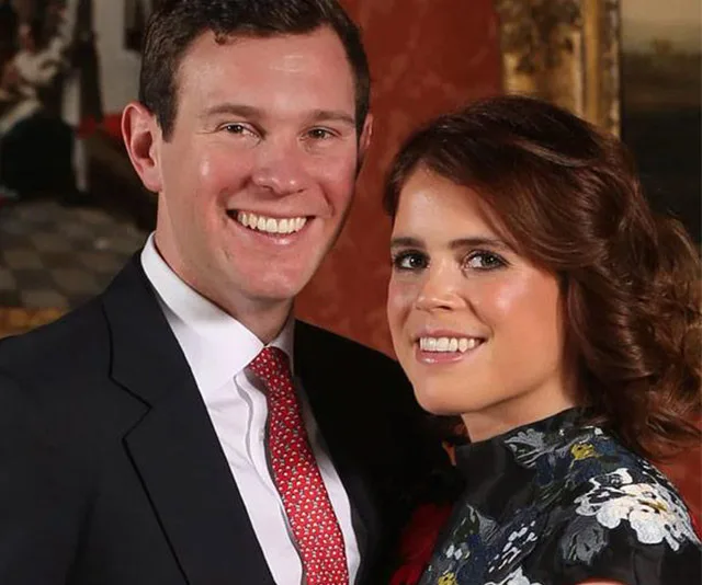 A smiling couple poses together, the woman in a floral dress and the man in a suit with a red tie, indoors.