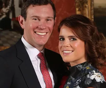 A smiling couple poses together, the woman in a floral dress and the man in a suit with a red tie, indoors.