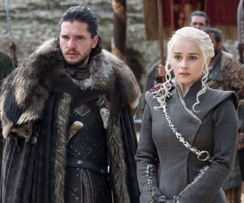 Two characters in medieval attire, one with dark hair and a fur coat, the other with braided platinum hair and a grey outfit.