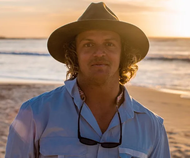 A man with curly hair and a hat stands on a beach during sunset, wearing a light-colored shirt with sunglasses hanging on it.