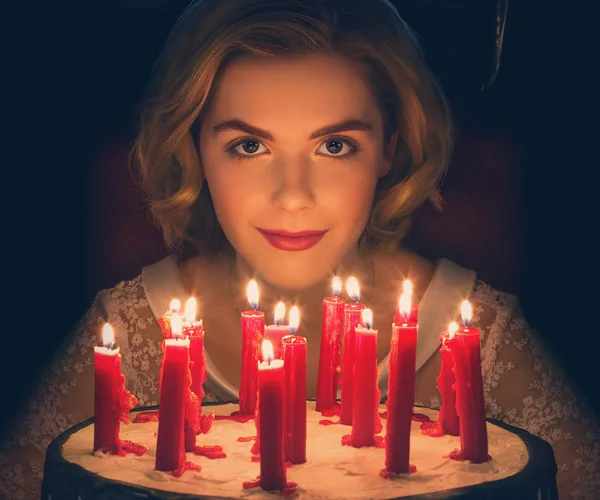 Young woman with blonde hair surrounded by lit red candles, wearing a lace dress in dim lighting, from "Chilling Adventures of Sabrina".