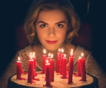 Young woman with blonde hair surrounded by lit red candles, wearing a lace dress in dim lighting, from "Chilling Adventures of Sabrina".