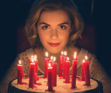 Young woman with blonde hair surrounded by lit red candles, wearing a lace dress in dim lighting, from "Chilling Adventures of Sabrina".