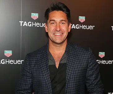 Person smiling at a TAG Heuer event, wearing a checkered blazer and black shirt, standing in front of a branded backdrop.