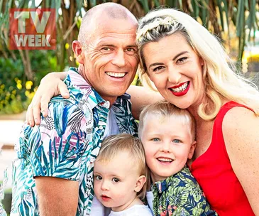 A happy family of four, with two adults and two young children, smiling outdoors in bright, patterned clothing.