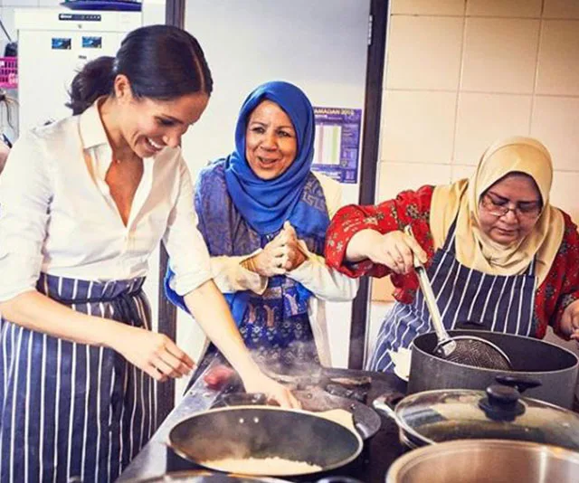 Woman in a white shirt cooking with two women in headscarves, all smiling, in a kitchen setting.