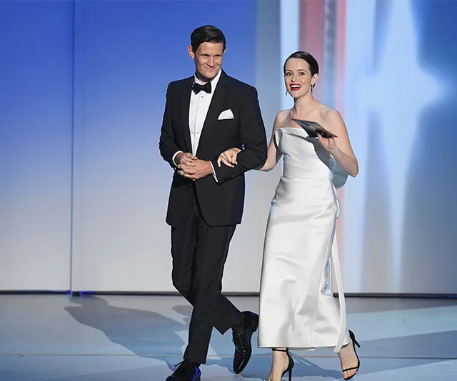 Two actors, one in a tuxedo and the other in a white gown, walk onstage at the 2018 Emmys, smiling and holding an award envelope.
