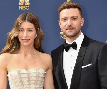 Jessica Biel in a strapless white gown and guest in a tuxedo at the 2018 Emmys, with an NBC logo in the background.