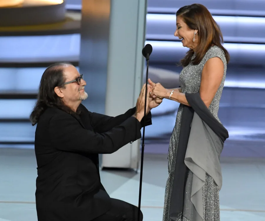 Man kneeling and proposing to smiling woman on stage, both dressed formally, during an award show.