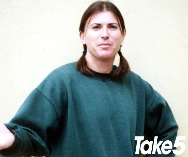A woman in a green sweater with pigtails stands with arms slightly raised against a plain background.
