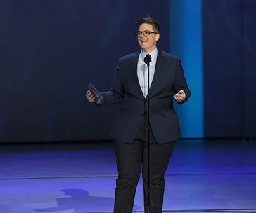 Hannah Gadsby on stage at the Emmys, wearing a suit, speaking into a microphone while holding an envelope.