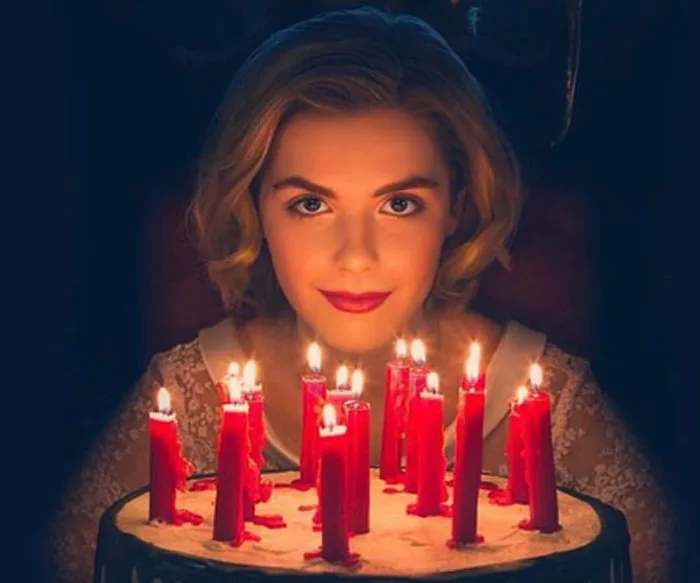 Young woman with blonde hair behind a cake lit with red candles, from "Chilling Adventures of Sabrina."